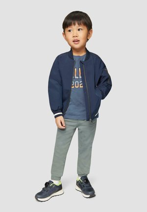 Marineblaues Bomberjackett mit weißen Streifenakzenten, graues T-Shirt mit Druck "2022", hellgraue Hose und dunkle Sneakers mit neongrünen Details.