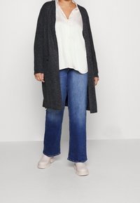 Dlouhý tmavě šedý kardigan přes světlou blůzu, v kombinaci s modrými džínami s širokými nohavicemi a béžovými botami. Kardigan má otevřený přední díl a dlouhé rukávy.