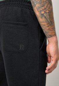 Tatoveret arm hviler ved siden af sorte bukser med elastisk talje og en lille lomme med et subtilt præget "JP π" logo.