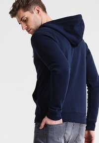 Sudadera azul marino hecha de tela suave, con capucha con cordón, mangas largas y puños de canalé, vista desde un ligero ángulo trasero.