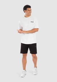 Vit kortärmad t-shirt med svart logotyp, tillsammans med svarta shorts. Modellen bär vita sportskor, står och klappar händerna.