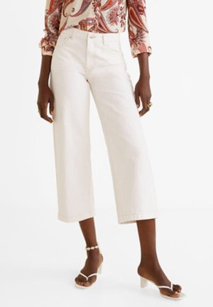 Personne portant un pantalon blanc large et court, un haut à manches longues à motifs, des sandales blanches à talons et un bracelet de cheville en perles.