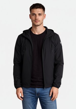 Młody mężczyzna ubrany w czarną kurtkę z kapturem i zamkiem błyskawicznym, pod którą ma czarny t-shirt oraz niebieskie dżinsy, stoi na jednolitym białym tle.