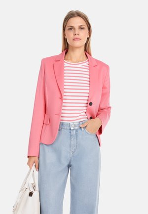 Rosa taillierter Blazer mit zwei Knöpfen, kombiniert mit einem gestreiften Oberteil in Weiß und Pink sowie hellblauen, hoch geschnittenen Jeans. Hält eine weiße Handtasche.