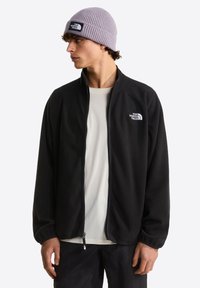 Μαύρο φλις μπουφάν με ψηλό γιακά και πλήρες φερμουάρ, συνδυασμένο με ανοιχτό γκρι ριμπ σκουφάκι με ετικέτα λογότυπου. Φορεμένο πάνω από λευκό πουκάμισο.