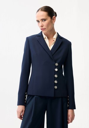 Kvinde med glatstrøget hår iført navyblå blazer med fem sølvknapper og matchende viddebenbukser mod hvid baggrund.