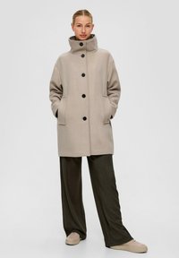 Beige wollen mix jas met een hoge kraag, vijf zwarte knopen en zijzakken. Gepaard met losse, donkergroene broeken en lichtgekleurde schoenen.