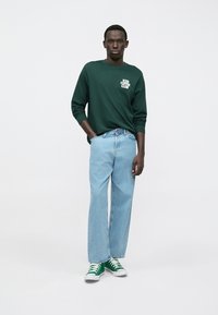 Zaļš krekls ar garām piedurknēm un uzrakstu "ICED COFFEE CLUB", kopā ar gaiši zilām taisnām džinsa biksēm un zaļiem sporta apaviem. Vienkāršs, ikdienišķs tērps.