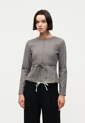 Rövid, sötét hajú nő, aki szürke, hosszú ujjú, testhez simuló felsőt visel elöl kötéssel, valamint fekete nadrágot fehér zsinórral, egyszerű háttér előtt.