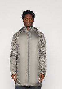 Hosszú, kapucnis lepedődzseki fényes, fémes szürke anyagból. Elülső cipzárral és enyhén elasztikus mandzsettákkal rendelkezik. Simább, könnyű tapintású.