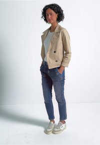 Beige doppeltknopfjacke, blaue jeans mit seitentaschen und graue sneakers mit hellen akzenten. Das outfit ist über einem weißen oberteil geschichtet.