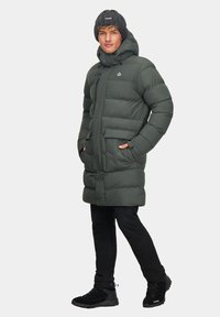 Ciemnozielona kurtka puchowa z kapturem, z przednim zamkiem błyskawicznym, dwoma bocznymi kieszeniami oraz czarną, prążkowaną czapką beanie. Całość uzupełniają czarne spodnie i buty.
