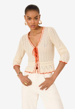 Woman wearing cream crochet cardigan with orange trim and tie front, paired with cream pants, gold hoop earrings, and bracelets.