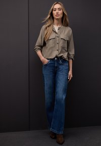 Chemise kaki à boutons avec deux poches, associée à un jean bleu à jambes larges et des bottines marron, sur un fond sombre.