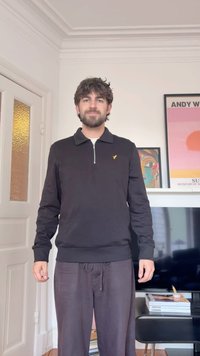 Homme aux cheveux foncés et à la barbe portant un pull à fermeture éclair noir et un pantalon ample foncé, debout dans un salon avec des œuvres d'art et une télévision derrière lui.