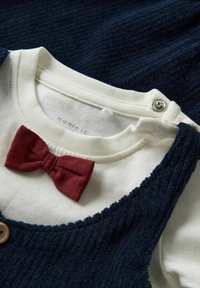 Vestido azul marino texturizado con una camiseta interior blanca, que presenta una pajarita de tela roja y un cierre a presión. El vestido tiene botones de madera y bordes deshilachados.