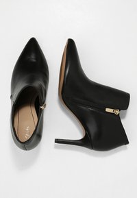 Stivaletti alla caviglia in pelle nera con punte appuntite, tacchi alti e cerniere laterali con dettagli in oro, texture liscia e design minimalista.