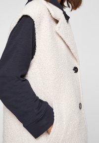 Gilet beige chiaro, tessuto strutturato con colletto, caratterizzato da due bottoni neri e tasche laterali, indossato sopra un top scuro a maniche lunghe.
