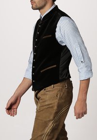 Schwarze Samtweste mit Satinakzenten, ausgestattet mit bronzenen Knöpfen, Seitentaschen und getragen über einem karierten blauen Hemd. Braune Hose rundet den Look ab.