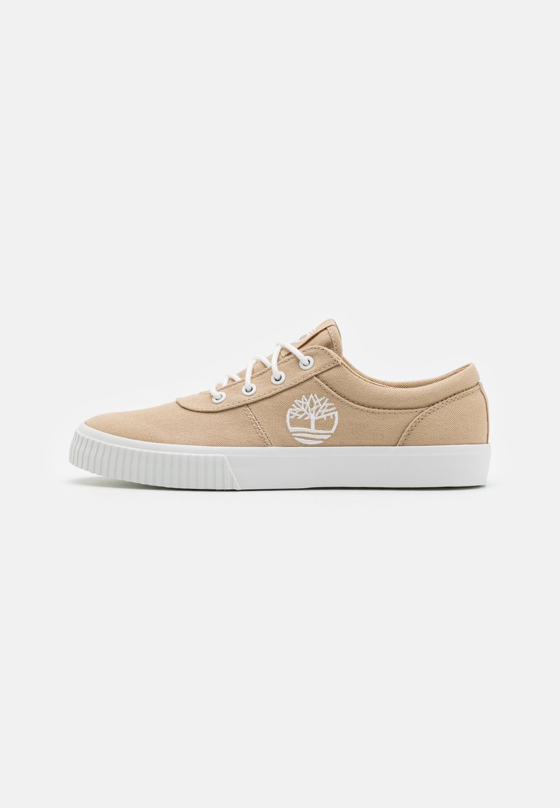 Beige canvas sneakers med vit gummisula, med vita ögla och snören, samt en vit logotypdesign på utsidan.