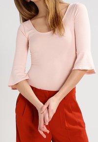 Světle růžový přiléhavý top s kulatým výstřihem a tříčtvrtečními zvonovými rukávy, skvěle ladící s červenými kalhotami s vysokým pasem. Hladká textura.