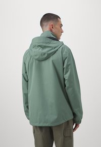 Zaļš kapuci žakets ar gludu, matētu tekstūru, elastīgām aprocēm un šņorējamu kapuci. Valkāts virs khaki krāsas cargo biksēm.