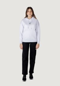 Sudadera blanca con capucha, bolsillo frontal y detalle de logo, combinada con pantalones negros y zapatos negros. Corte recto y ajuste casual.