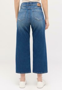 Jean bleu à jambes larges en denim ; présente une taille haute, deux poches arrière et un patch en cuir avec le logo sur la ceinture.