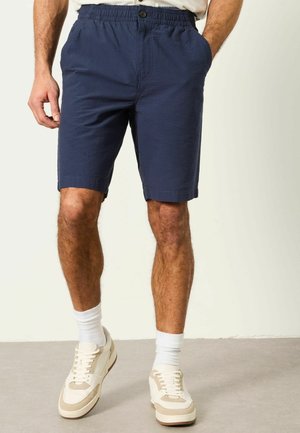 Mann trägt marineblaue, strukturierte Shorts mit elastischem Bund und Knopf, weiße Socken und weiße Sneakers, steht auf grauem Boden.