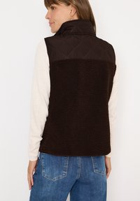 Chaleco marrón acolchado con un cuello alto, que presenta una parte trasera de forro polar suave y texturizado. Combinado con una camiseta de manga larga ligera y jeans azules.