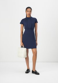 Mörkblå klänning med kort ärm, en åtsittande bodice och något utställd kjol. En texturerad vit handväska kompletterar outfiten, som bärs med svarta loafers.