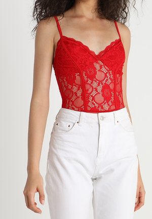 Femme portant un haut sans manches en dentelle rouge avec des fines bretelles et un pantalon blanc taille haute sur fond uni.