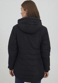 Svart pufferjakke med hette, quiltet design, elastiske mansjetter og buet søm. Stoffet ser glatt og litt glansfullt ut.
