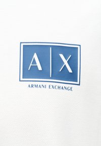 Μπλε ορθογώνιο με λευκά γράμματα "A | X" πάνω από μπλε κείμενο "Armani Exchange" σε υφασμάτινο φόντο με υφή λευκού χρώματος.