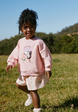 Lille barn med krøllet hår iført en pink sweater med zebra-design og matchende nederdel går udendørs på en græsmark under klar himmel.