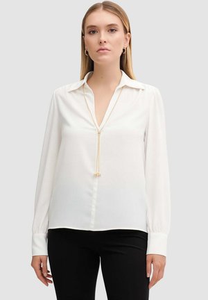 Donna che indossa una camicetta bianca a maniche lunghe con colletto, pantaloni neri, orecchini a cerchio dorati e una lunga collana d'oro su uno sfondo grigio.