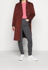 Lungo cappotto bordeaux con un unico bottone, abbinato a una t-shirt rosa e jeans grigi. Sneakers bianche completano l'outfit. Tessuto morbido e testurizzato.