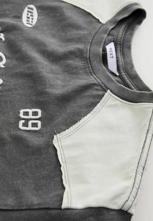 Grauer Sweatshirt mit weißen Schulterakzenten, mit gesticktem Text und Zahlen. Das Material wirkt weich und hat ein lässiges Design.