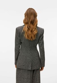 Hallhallane tweed bleiser, millel on struktureeritud õlad, kohandatud istuvus ja taga vent. Kanga pinnal on elegantne tekstuur, mis lisab sügavust.