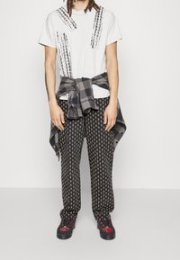 Persona con una camiseta blanca con estampados de huellas de neumáticos negros, pantalones a rayas en negro y gris, una camisa de cuadros atada a la cintura y zapatillas negras con detalles rojos.