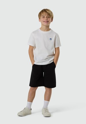 Garçon blond souriant, debout les mains dans les poches, vêtu d'un t-shirt blanc, d'un short noir, de chaussettes blanches et de baskets blanches sur un fond uni.