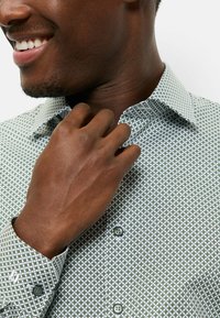 Weißes Hemd mit geometrischem Muster in Grün und Marineblau, ausgestattet mit Kragen und geknöpften Manschetten. Der Stoff wirkt glatt und leicht.