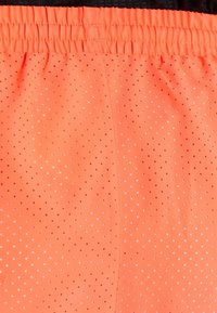 Pomarańczowe szorty sportowe o siateczkowej strukturze, z małymi perforacjami na całej powierzchni i elastycznym paskiem w talii, umożliwiającym regulację dopasowania.