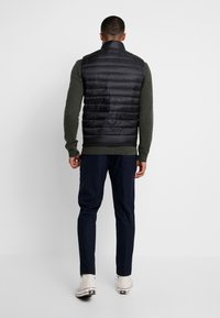 Czarna pikowana kamizelka z poziomym wzorem, wysokim kołnierzem i prążkowanym dołem, noszona na zielonym swetrze o fakturze, zestawiona z granatowymi spodniami i białymi butami.