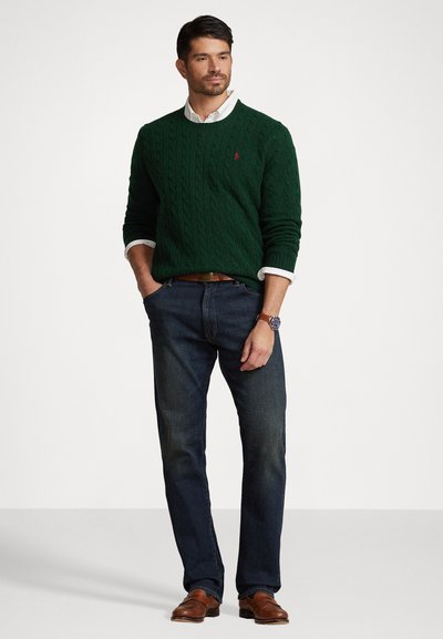 Suéter verde de punto trenzado, jeans azules, zapatos de cuero marrón y un reloj. El suéter tiene un escote redondo y puños acanalados.