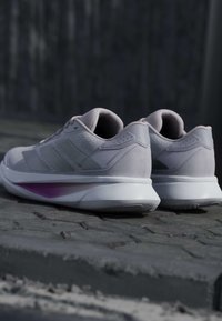 Hellgraue Sportschuhe mit weißen Zwischensohlen und dezenten lila Akzenten, die auf Kopfsteinpflaster liegen.