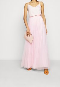 Ljusrosa tulle-maxikjol med ett figurnära midjeband, kombinerad med en satin-camisole; bär en liten rosa clutch; orange sandaler med remmar.