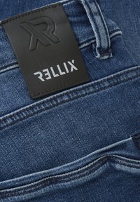 Donkerblauwe spijkerbroek van denim met een getextureerd zwart leren etiket waarop "RELLIX" in zilver is gestempeld, en zichtbare stiksels langs de tailleband.