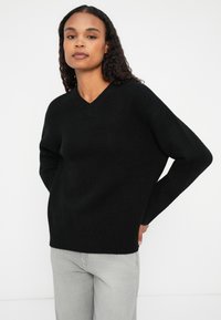 Svart v-hals genser med lange ermer, laget av strukturert strikket stoff. Modellen har på seg lysegrå jeans. Enkel og minimalistisk design.