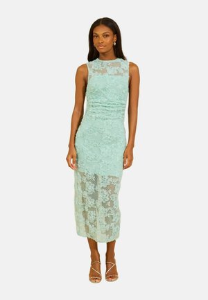 Kobieta ubrana w bezrękawową, jasnozieloną koronkową sukienkę midi z przezroczystym kwiatowym wzorem i cielistymi sandałkami na paskach, stojąca na białym tle.